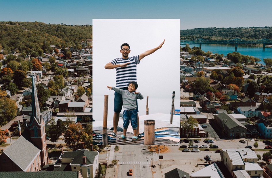 This image shows a man and a child posing in front of a white background, which is digitally superimposed over an aerial view of a town near a body of water.